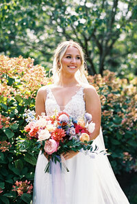 Bride smiling in garden holding vibrant summer bouquet with pink, coral, and purple flowers at Gervasi Vineyard.