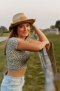 Image of senior girl leaning against a fence while holding the edge of her cowgirl hat and giving the camera a soft smile.