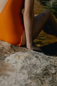 A stock photo of a woman in an orange bathing suit sitting with her legs in the water