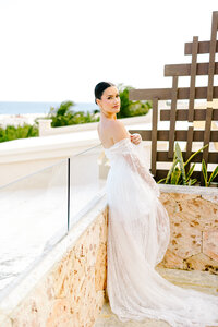 Blonde bride in a fitted lace gown and cathedral-length veil smiles over her shoulder while holding a tropical bouquet, walking through a sunlit corridor in Cabo San Lucas, Mexico.

