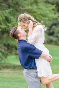 a man wearing a dark blue polo shirt and gray slacks lists up his fiancee wearing a while lace dress as they stand in a grass field in American Fork. Captured by Salt Lake City Photographer Melissa Woodruff Photography