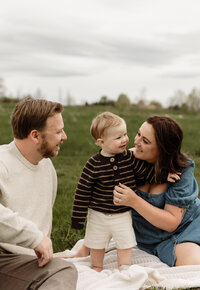 A family of three relaxes in a scenic field, captured beautifully by a New England photographer. The father gently kisses their baby’s head as the mother rests on his lap, cradling the little one on her tummy. Dressed in soft, timeless tones, they’re surrounded by lush greenery — a tender, fine art moment filled with warmth and connection.