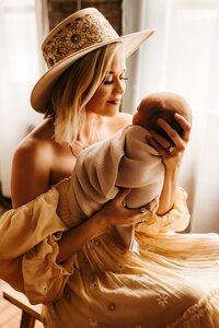 A woman wearing a floral hat is holding her newborn baby and gazing into their eyes during her newborn photoshoot captured by Infinite Productions