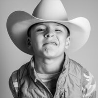A young boy wearing a cowboy hat and a quilted vest makes a playful face, scrunching his nose and lips together in a humorous expression.