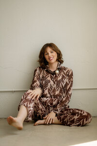 A woman in a maroon and brown jumpsuit sits on the floor and leans against the wall