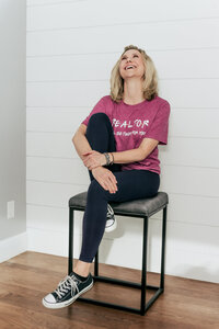 Kristin in a casual pink top and black pants, seated on a modern stool with playful pose