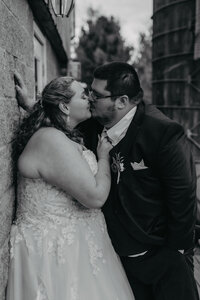 black and white portrait of bride pulling groom's tie to bring him in for a kiss while leaning against a wall