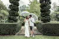 A bride and groom standing in a garden, holding umbrellas and smiling at each other. The bride is wearing a white wedding gown while the groom is wearing a white polo shirt.