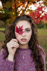 A curly haired blue eyed high school senior girl laying in the grass surrounded by fallen red Maple leaves. Captured by Springfield, MO photographer Dynae Levingston