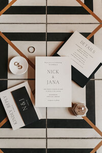 Minimalist black and white wedding invitation suite with gold geometric accents styled in a flat lay on a modern striped surface, perfect for elegant and contemporary wedding stationery inspiration.