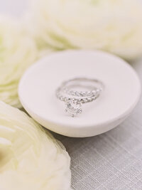 Elegant close-up of diamond engagement ring and wedding bands styled on a white ceramic dish — fine-art wedding details captured by Asia Pimentel Photography.