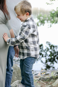 Toddler boy in a plaid shirt touching his mom's pregnant belly during a NH Family Photographer session