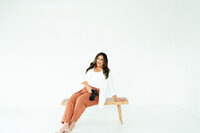 Adriana Montoya Photo sitting on a small bench, her ankles crossed. Her arms are behind her, supporting her as she sites. Her camera is in her other hand.
