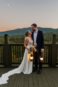 A bride and groom stand on a hiking trail surrounded by lush green pine trees with a waterfall off in the distant background. The bride is in a flowy wedding dress with a long veil and the groom is wearing tan pants with an orange leather jacket. 