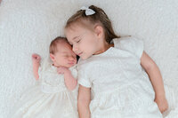 Newborn baby and big sister wearing white heirloom dresses snuggled up on a fluffy blanket with their eyes closed.