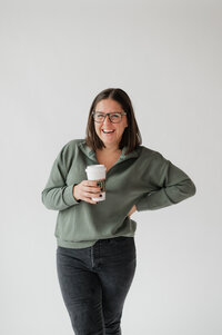 A person smiling with one hand on her hip and the other holding a coffee cup. 