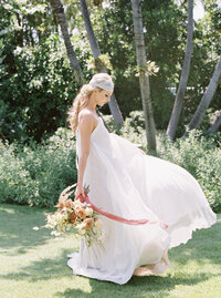 A woman in a wedding dress with a white head wrap sways holding down a lush bouquet.