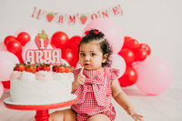 baby girl sitting on floor with birthday cake with red balloons in background