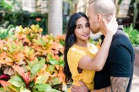 A black man in baltimore kisses the forehead of his Indian fiance who is wearing bright yellow. She smiles at the camera , surrounded by orange leaves