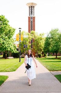 Senior walking with her cap in hand down the sidewalk of Belmont University bell tower in the background
