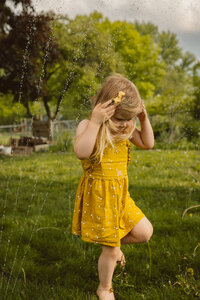 toddler girl wearing yellow sun dress and yellow bows in her hair emerges from a sprinkler while playing in the backyard