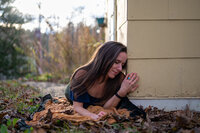 A woman rests against the outside of a yellow house in a self-portrait, surrounded by fallen leaves, capturing introspection and creative storytelling.