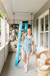 Lady diyer carries a ladder on her front porch.