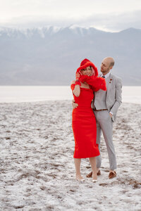 A woman in red smiles at the camera while a man next to her has his arm around her waist and looks at her