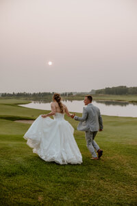 Bride and groom in wedding attire walk on a grassy field at sunset, near a tranquil lake. The sky is softly lit, creating a serene, romantic mood.