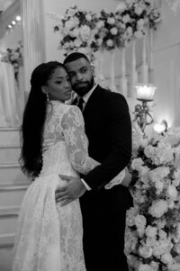 A couple embraces lovingly in a black-and-white photo. The woman wears a lace dress, the man a suit. They are surrounded by flowers, creating a romantic mood.
