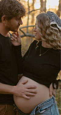 A husband holds his pregnant wife's belly while she holds his chin.