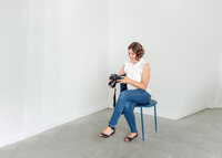 Calgary Brand Photographer Kate Daniel Photography sitting in a white room on a blue chair, wearing blue jeans and a white shirt, looking down at her camera.