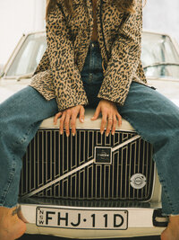 Person in a leopard print jacket and jeans sits on the hood of a white Volvo car with license plate FHJ-11D, capturing the bold style and brand design inspiration behind Sweet Pea Ink Creative.