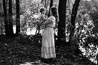 Black and white photo of a mom holding a toddler while they smile at each other in a forested area.