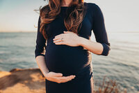 a woman holds her baby bump while wearing a navy blue dress on the beach at sunset cliffs in san diego