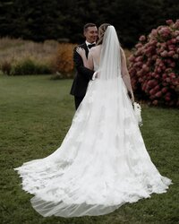A bride and groom stand on a grassy lawn, surrounded by lush greenery and colorful flowers. The bride wears a long white gown and veil, holding a bouquet, while the groom is in a black suit. Captured by a Fine Art Weddings &amp; Portraits New England photographer, they gaze at each other lovingly.