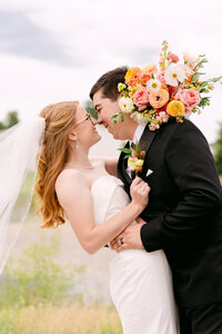 A bride and groom in formal attire share a close embrace and smile while touching noses, outdoors. The bride holds a colorful bouquet of flowers.