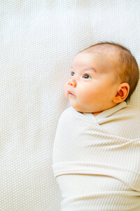 Austin Texas Photographer | A newborn baby swaddled in a beige blanket lays on a white bed with his eyes wide open.