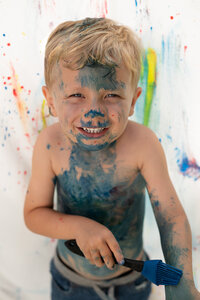 Young blonde boy smiling and covered  in blue paint in front  a painted surface