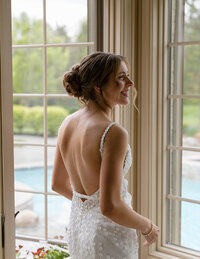 Bride in a floral lace gown stands by a window, looking to the side with a smile. Her hair is in an elegant updo, and a pool and garden are visible outside.