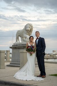 Newlywed couple posing for wedding photos next to ornate stone lion statue at the White Room venue in historic downtown Saint Augustine, with bride in white lace dress holding colorful bouquet and groom in navy suit with red tie, bridge visible in background under cloudy sky