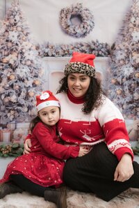 Mother and daughter dressed in matching red Christmas outfits and Santa hats, cuddling in front of a wintery holiday backdrop with frosted trees and a festive wreath.