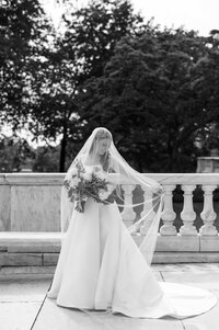 Black and white photo of a bride in a flowing gown holding her bouquet, gracefully lifting her veil in an elegant outdoor setting with a stone balustrade.