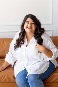 Marissa Martinez wearing a white top and blue jeans sitting on a brown leather couch