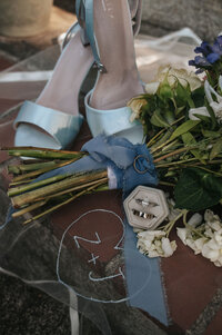 Light blue wedding shoes beside a colorful bridal bouquet, wedding rings, and a blue embroidered heart with couple’s initials, captured by a Portland wedding photographer.