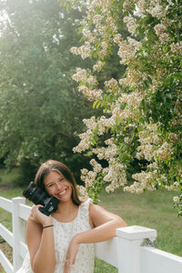 Portrait of Esperanza, lead photographer at EC Photography, smiling while holding a camera near a white fence in Alabama