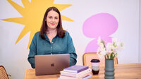Claire Dicarlo sitting in front of a colourful wall, looking friendly while sitting at a laptop.