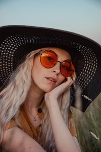 Amber in a field at golden hour wearing a black straw hat and round orange sunglasses