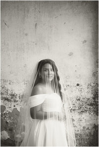 A bride stands against a rustic wall with her vail pulled over her smiling in black and white