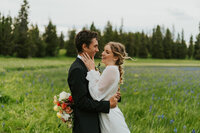 Bridal shoot in Rexburg, Idaho. A groom is holding his bride by the waist and looking at her. The bride is holding his neck with one hand and her wedding bouquet in the other behind his back. They're outside in a long grass field with pine trees in the distance.